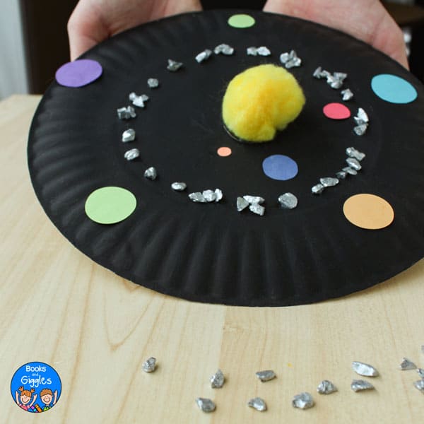 paper plate painted black with a yellow pom pom, construction paper circles and pebbles painted silver on top