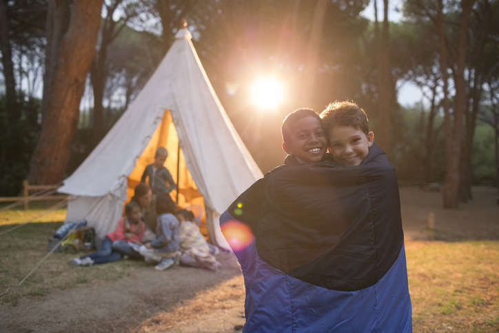 Two children stand together in a sleeping bag in front of a tent.