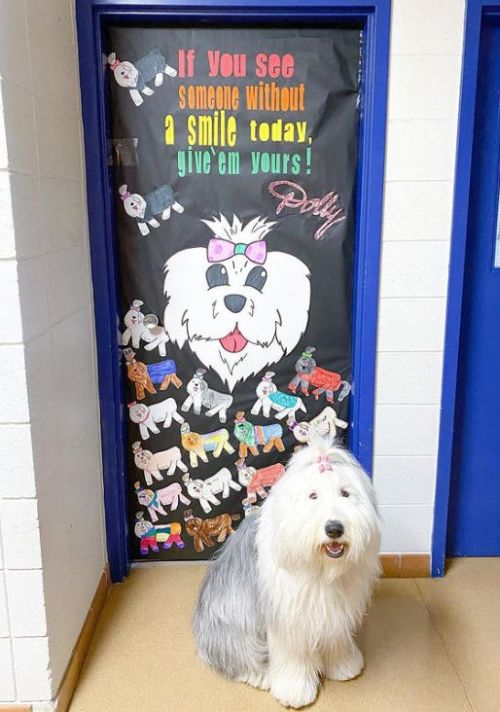 Dog standing by a classroom door decorated with smiling dogs. Text reads If you see someone without a smile today, give them yours.