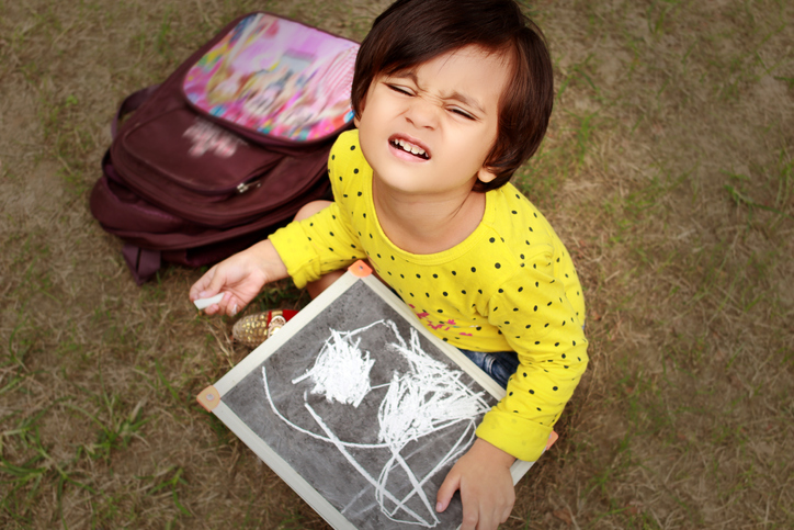 Drawing games can involve closed eyes drawing like this one pictured. A little girl sits with eyes closed holding chalk in front of a chalk drawing.