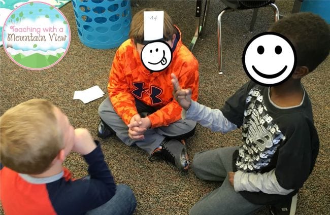 Fourth grade students sitting in a circle playing a math game, wearing cards on their heads with a number written on it
