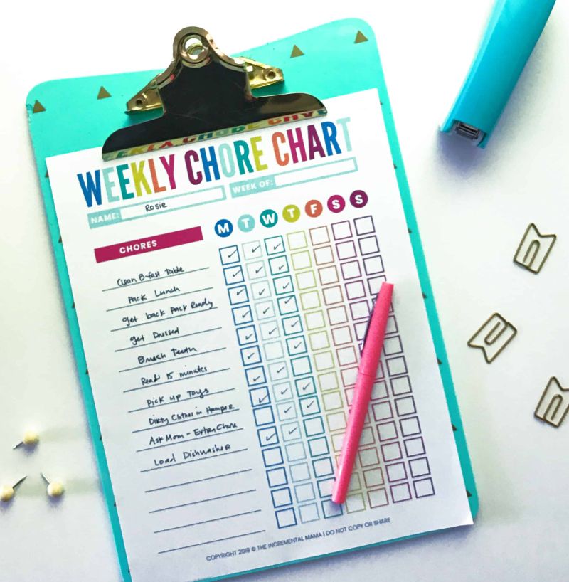 Blue clipboard holding a colorful chore chart checklist for kids, with a pen and other office supplies