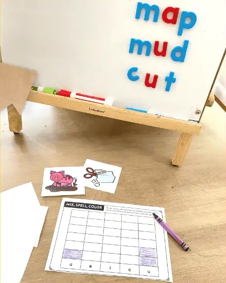 pictures, crayons, and a worksheet with a few squares colored in and magnetic letters on a whiteboard in the background  