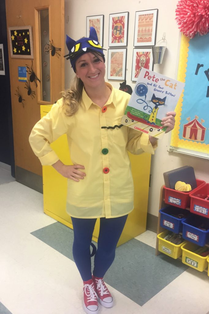 A woman is shown holding a Pete the Cat book. She is wearing a yellow button up shirt and signature red sneakers. She is wearing a pete the cat headpiece.