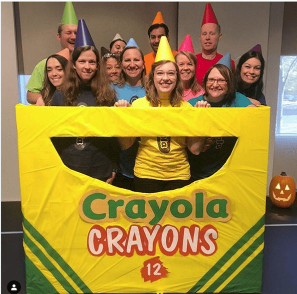 About a dozen people are wearing different colored pointy hats and different colored shirts. They are all standing inside a huge yellow box that says Crayola Crayons 12.