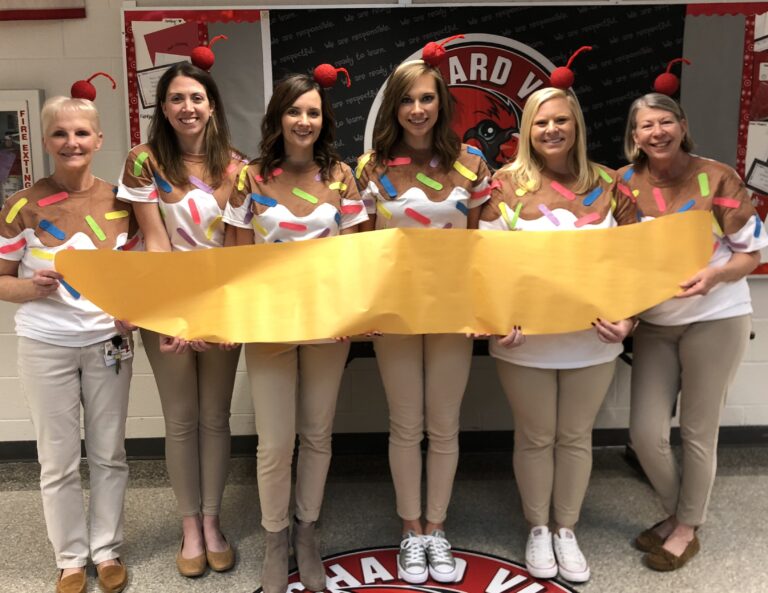 six teachers are shown with cherries on their heads wearing shirts with sprinkles on them. They are holding a large cut out of a banana.