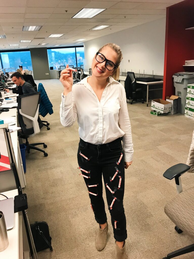 Teacher Halloween costumes can be simple like this one in which a woman is shown wearing a white collared shirt, glasses, and pants with smartie candies taped onto them.