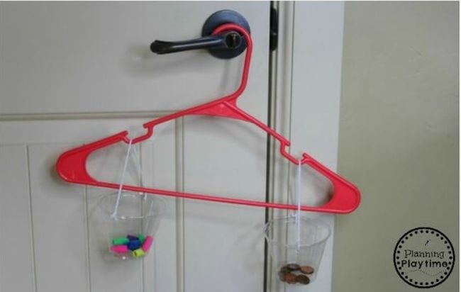 A red hanger hands from a doorknob. Hanging on each end is a plastic cup on a string with different items inside