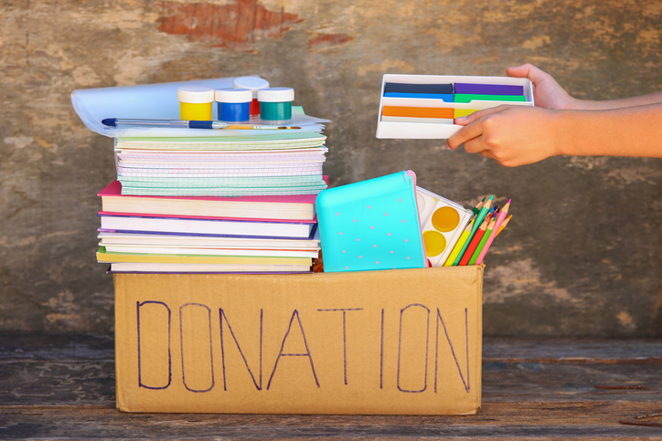A cardboard box has donation written on it. It is filled with school supplies.