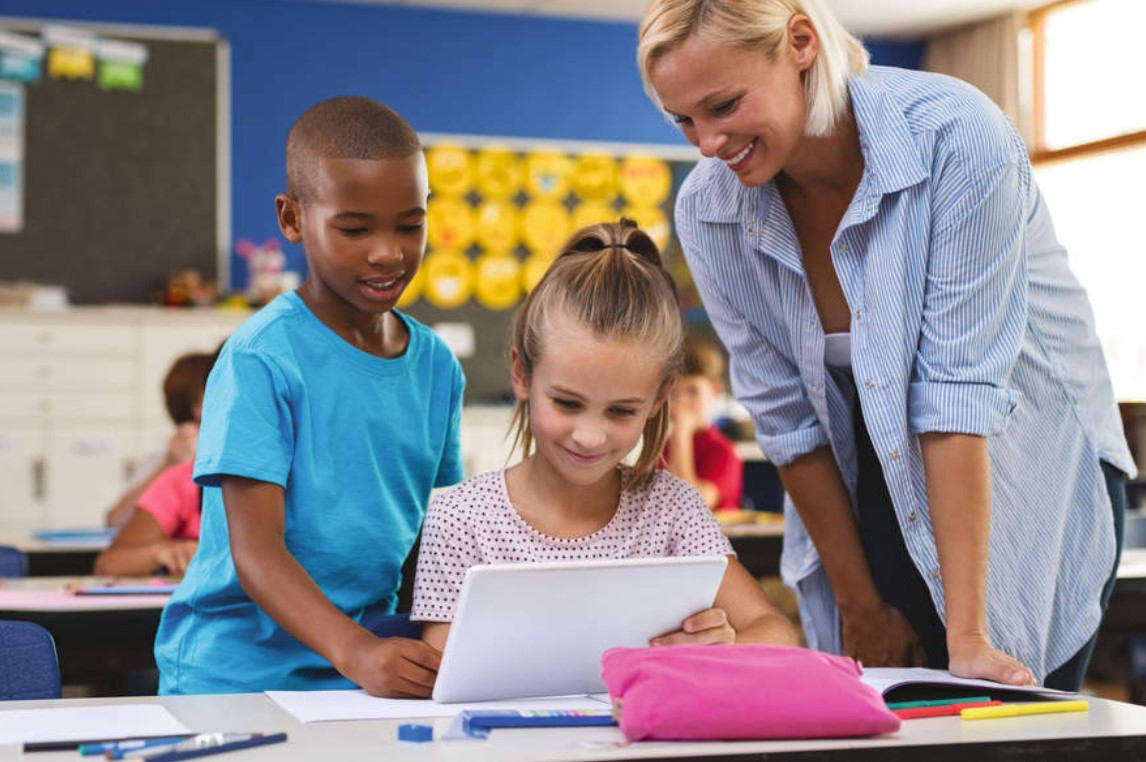 Multiethnic young students using digital tablet in classroom with teacher. Teacher in class with kids using electronic tablet.