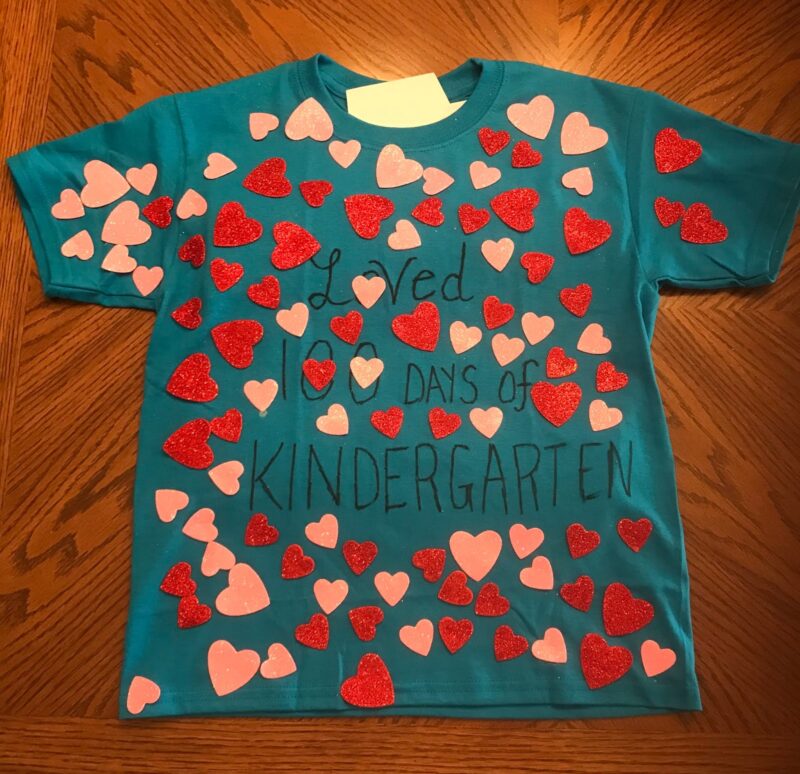 A teal t-shirt has red and pink heart stickers all over it. It says Loved 100 Days of Kindergarten in marker. (100th day of school shirt ideas)