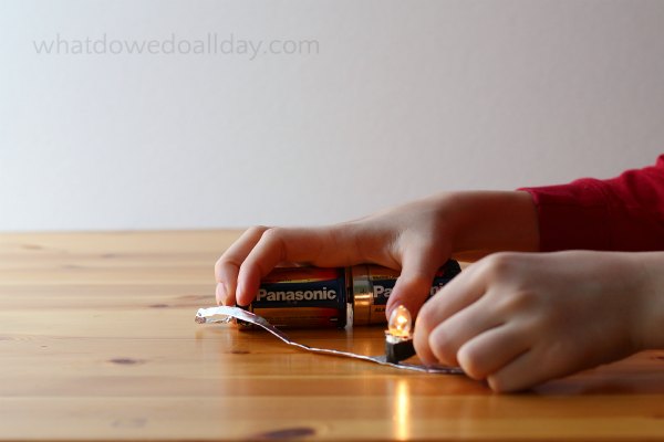 A child's hands are shown holding large batteries, tin foil, and a small light.