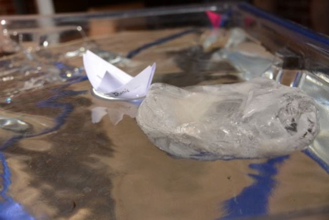 Paper boat floating in a dish with a large chunk of ice