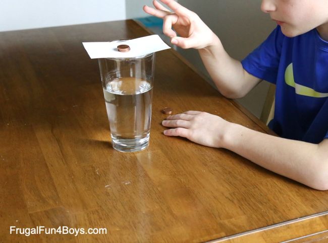 Third grade science student flicking an index card out from underneath a stack of pennies suspended over a cup of water