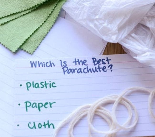 Card with text Which is the best parachute? Plastic, paper, cloth. Surrounded by pieces of fabric, plastic, and string.