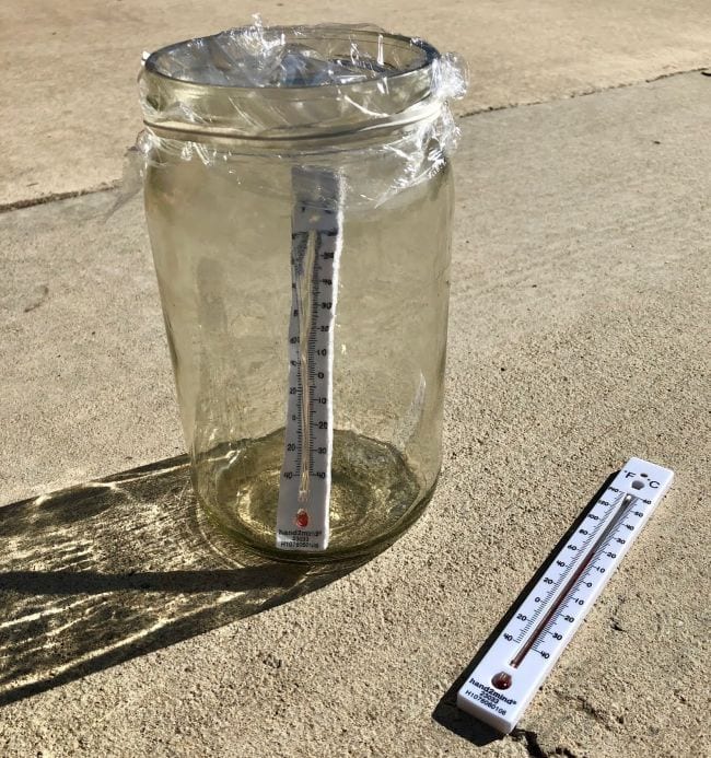 Glass jar with a thermometer inside, covered with plastic wrap, next to another thermometer lying on the sidewalk