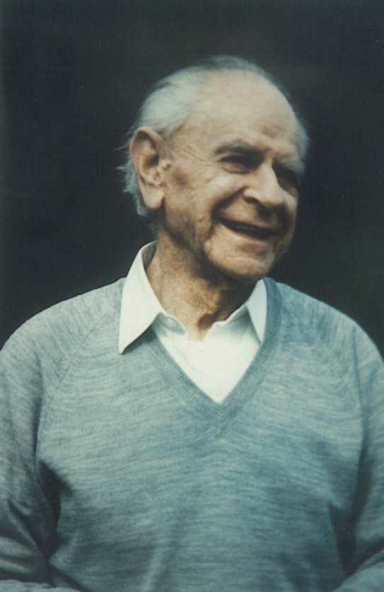 An older man is seen wearing a blue sweater with a white collared shirt under it. He is smiling with his head turned in a three-quarters view.