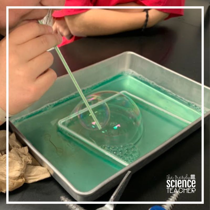 A student using a straw to blow a bubble inside another bubble in a pan of green liquid