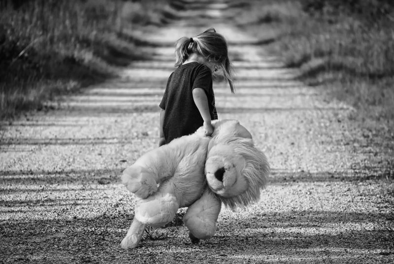 A little girl holding a very large teddy bear, dragging it down a dirt road