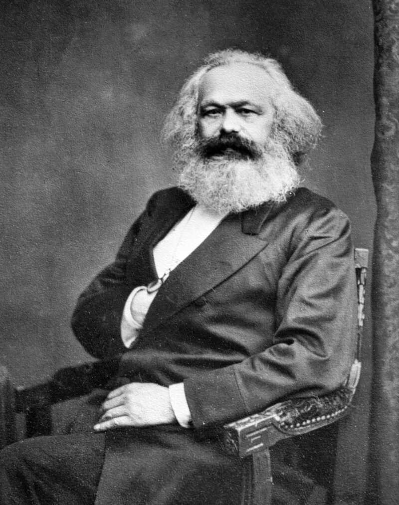 A black and white photograph of a seated man is shown. He has a large beard and his hand is in his suit breast pocket.