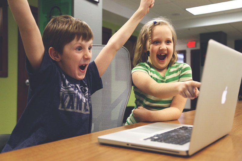 A young boy raises his arms in triumph as a young girl points at a computer screen, smiling (Picture Writing Prompts)