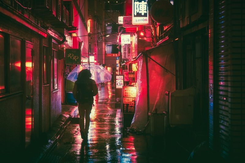 A person holding an umbrella walks down an alley toward a street filled with neon lights