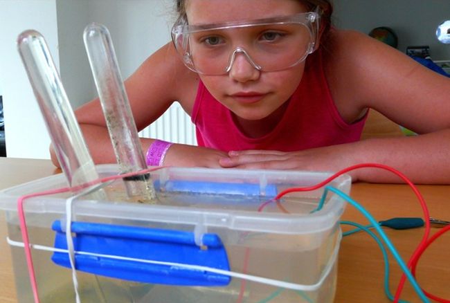 Eighth grade girl wearing goggles, looking at a container of water with test tubes and electric wires