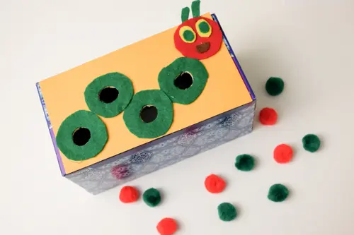 A tissue box has a caterpillar on the top of it. The body has holes in it. There are red and green pom poms to put inside the box in this example of very hungry caterpillar activities. 