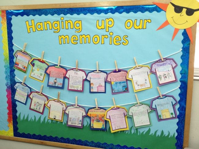 A bulletin board has a blue background with a sun in the corner. It has clotheslines on it with clothespins holding up t-shirts that students have written and drawn on. (summer bulletin boards ideas)