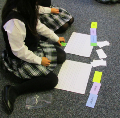 A girl in a school uniform sits on the carpeted classroom floor playing a grammar game
