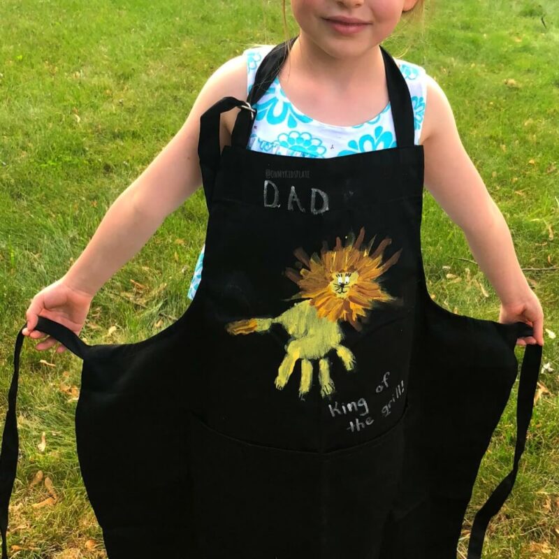 A child is seen wearing a black apron with a handprint design on it in this example of Father's Day crafts for kids.