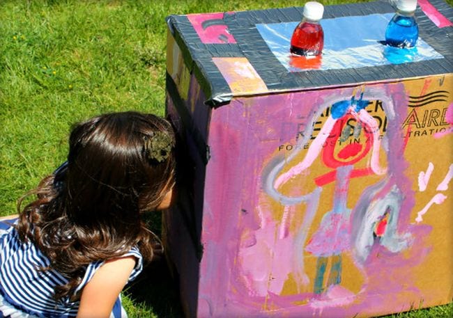 Child peering into a painted cardboard box turned into a light box with colored water in bottles
