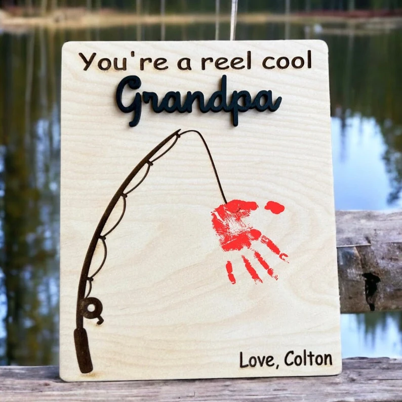 A wooden slab has You're a real cool Grandpa at the top and a fishing rod with a handprint on it. 