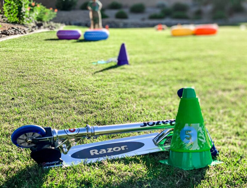 Some inflatable tires and cones are laid out in the background and in the foreground a folded Razor scooter is seen beside a green cone in this example of obstacle courses for kids.