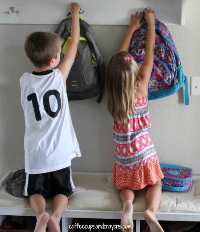 Two children hanging up their backpacks on hooks on the wall