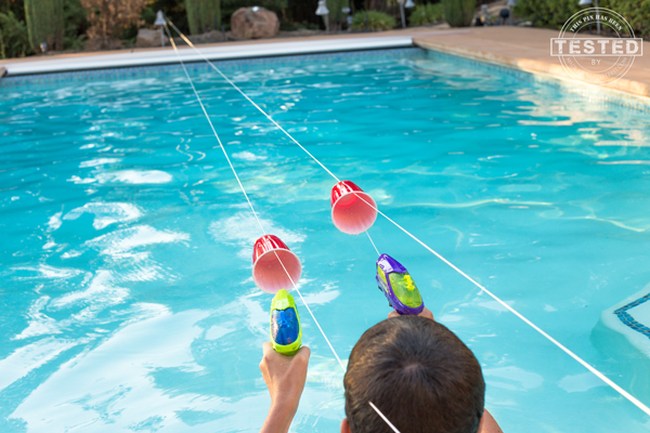 Kids using squirt guns to propel plastic cups along strings strung over a swimming pool
