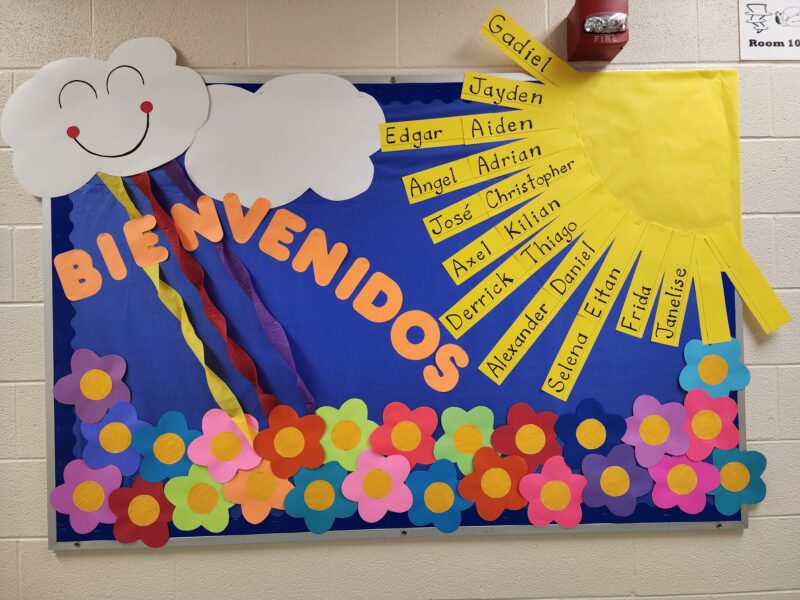 A cloud in the corner of a bulletin board has a smiley face on it. Flowers decorate the bottom of the bulletin board. A sun is in the right hand corner with a spanish and english name written on each ray.