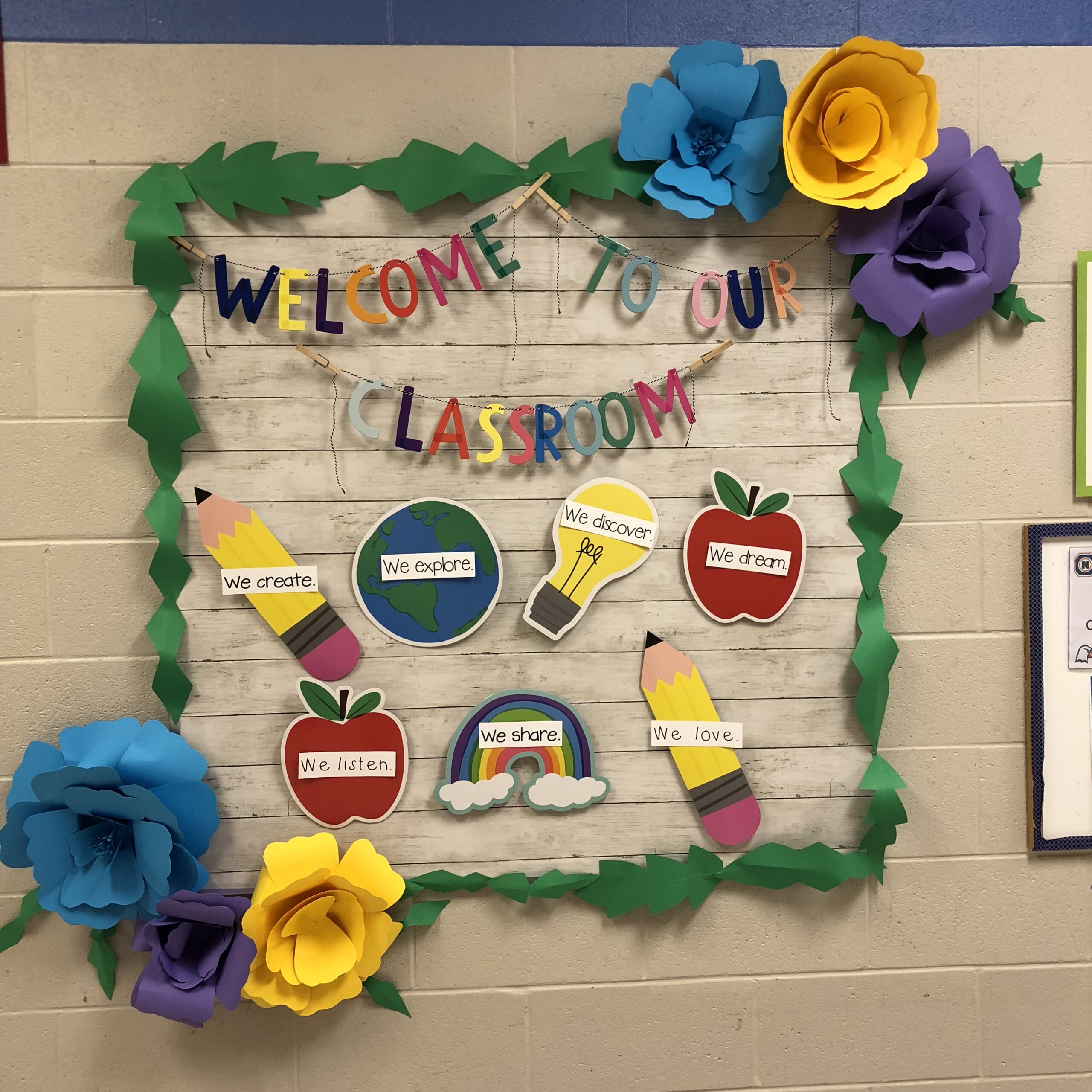 A sign says Welcome to Our Classroom. The corners of the bulletin board have large oversize 3D flowers.