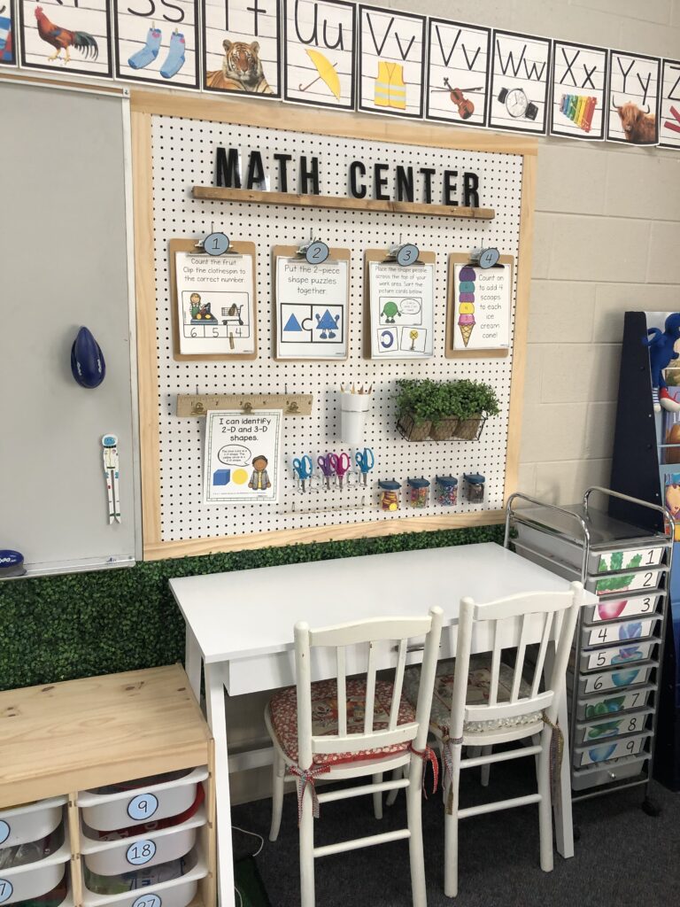 A corkboard says Math Center at the top and has several clipboards with different math assignments on it. A desk with two chairs is underneath it.