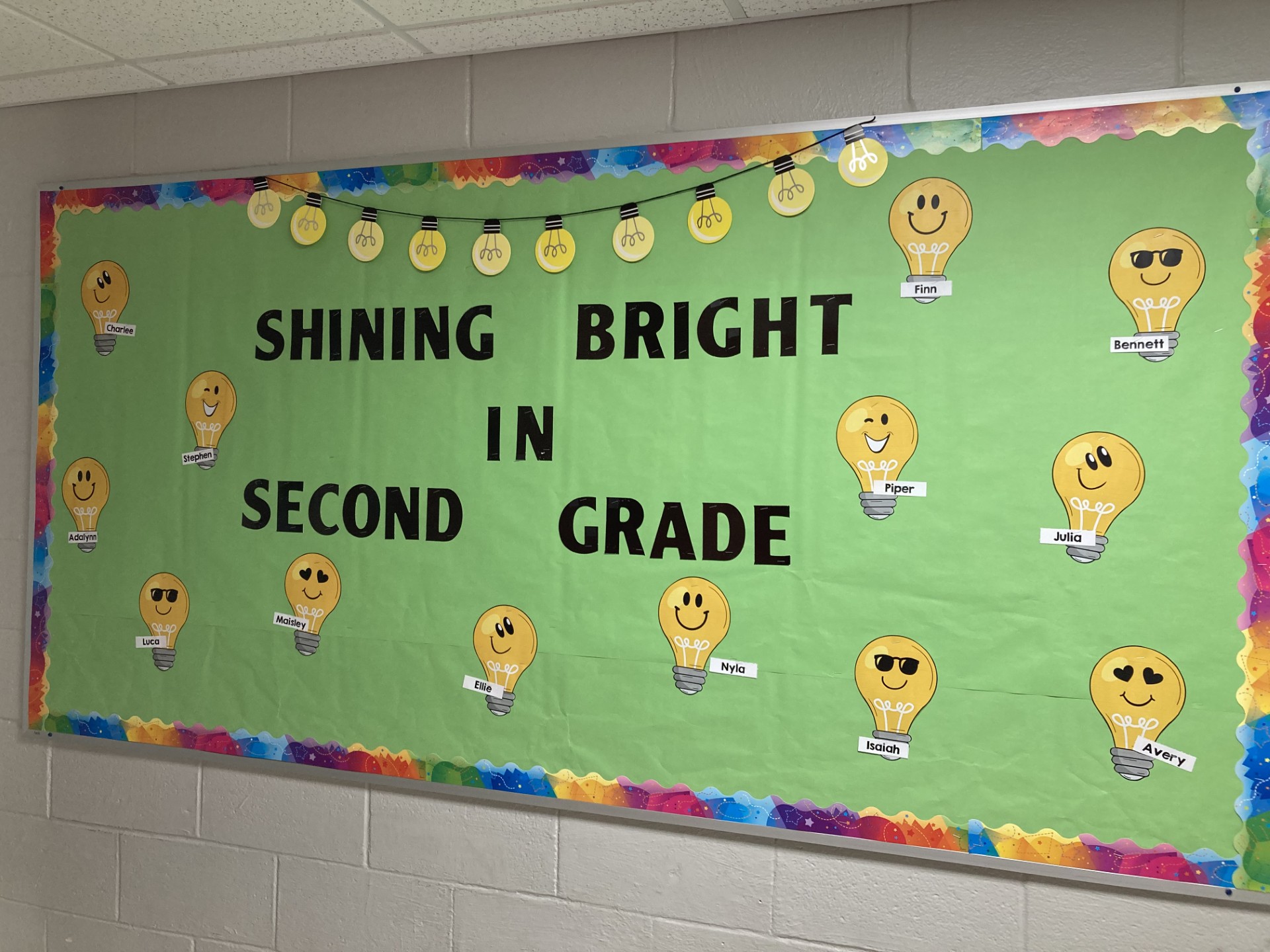 Little light bulbs with student names on them are featured. Bulletin board says Shining Bright in Second Grade.