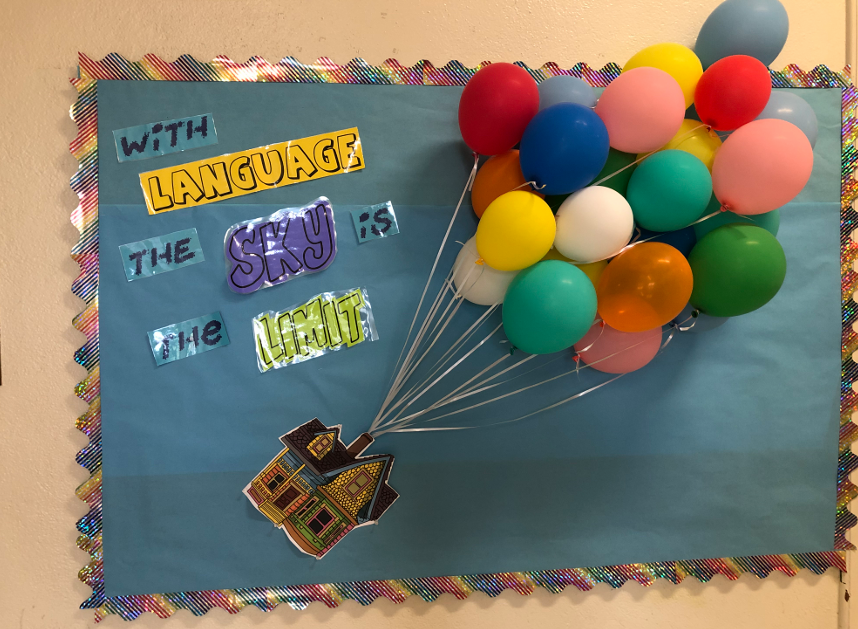A bulletin board has a small house on it that looks like it is being carried away by a bouquet of actual blown up balloons.