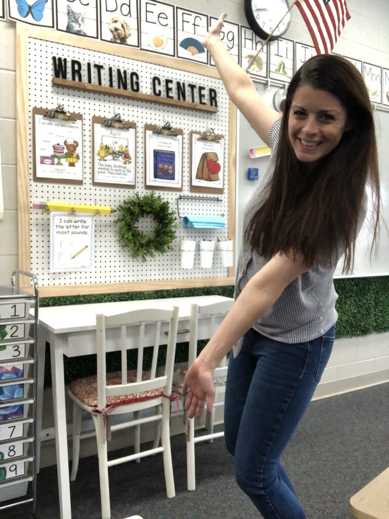 A teacher stands with her arms out showing off a board that says Writing Center. There are several clipboards attached to it.