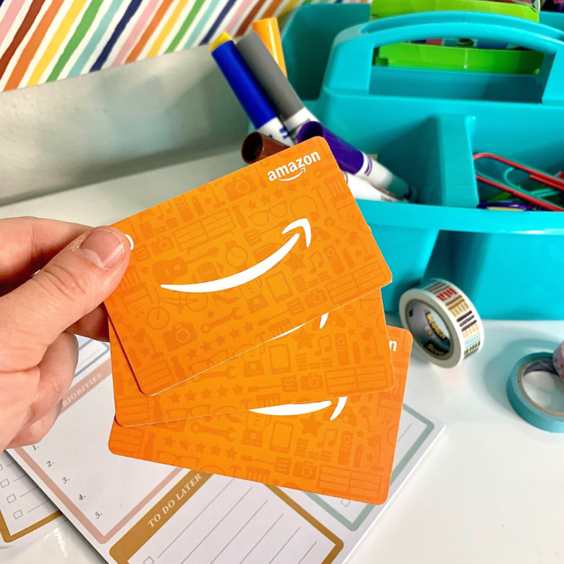 Hand holding two Amazon gift cards above a desk with art supplies.