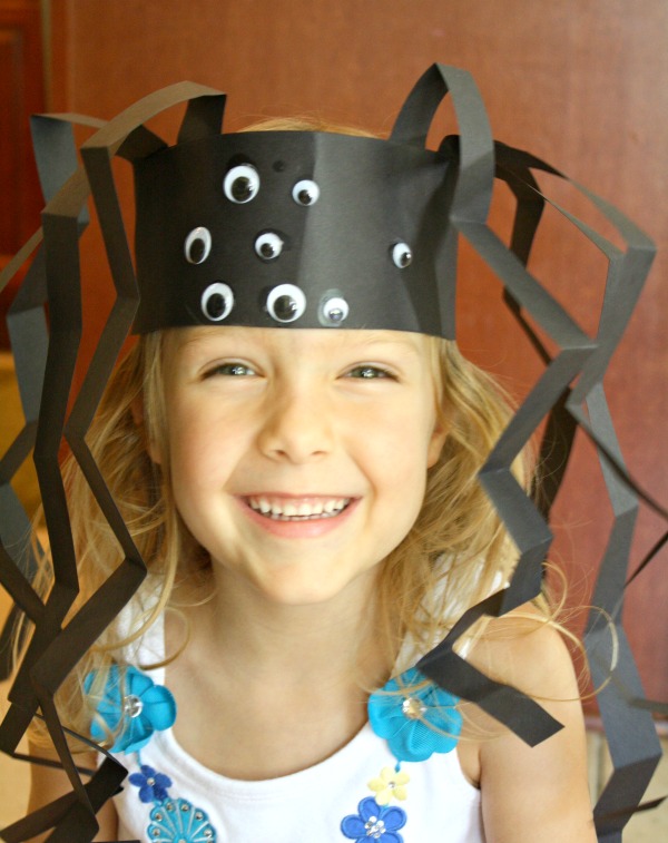 A smiling girl wears a spider headband made from black construction paper and many googly eyes