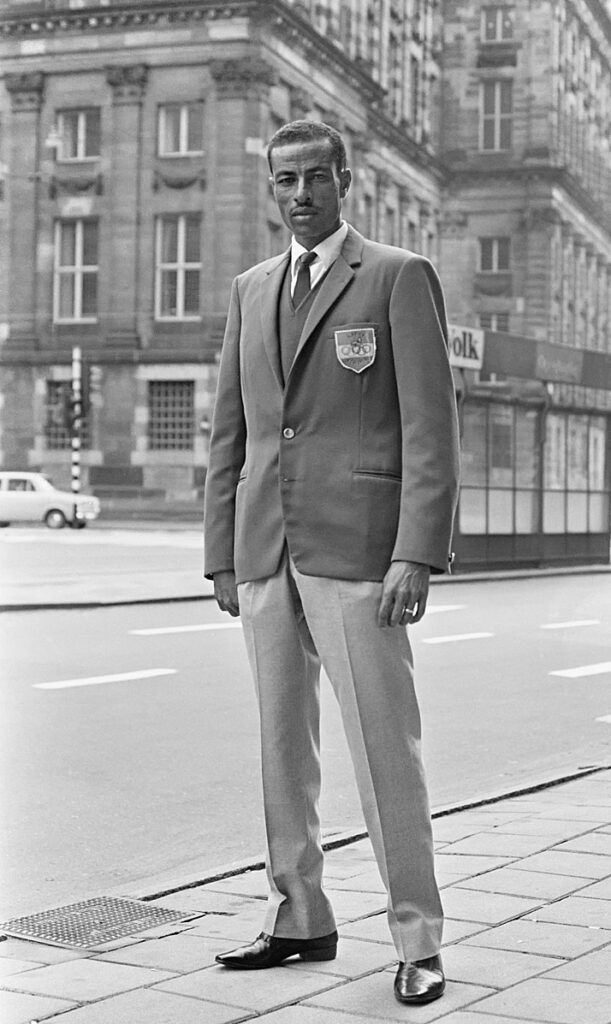 Abebe Bikila wearing suit standing in city street.