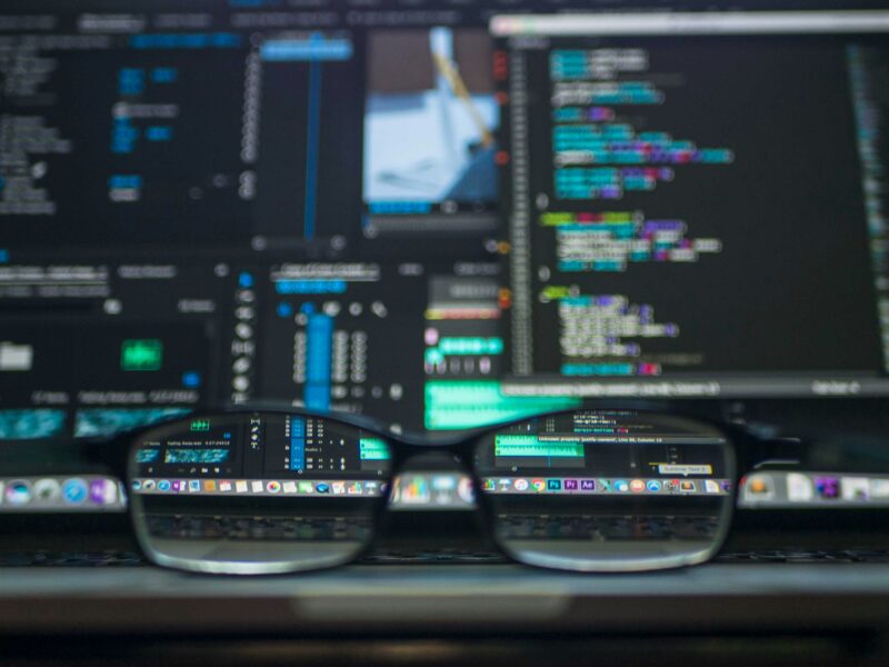 A pair of glasses sits in front of A collection of computer monitors displaying programming text