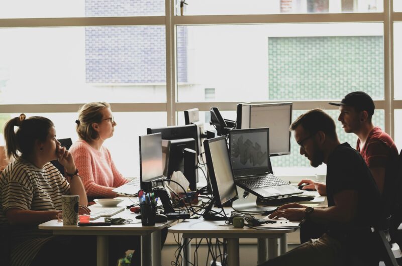 A group of DevOps programmers works at laptops around a table in front of a window