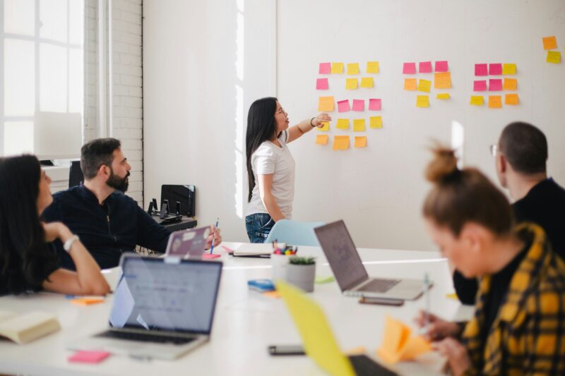 An IT project manager stands at a board of sticky notes as her team works at laptops around a table